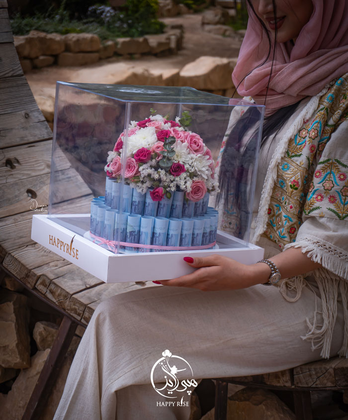 دختری ایرانی با لباس رنگ روشن روی صندلی واقع در طبیعت نشسته است که در دستانش هدیه دست ساز و شخصی سازی شده به نام کیک پول است که می توان آنرا در مناسبت های مختلف مانند تولد و روز ولنتاین هدیه داد.